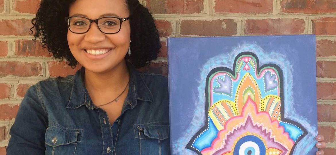 Female student Natalie Arthur stands holding her painting of a mandala