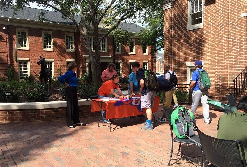 Folding table with orange tablecloth is set up in front of Belk Dining Hall with students gathered around